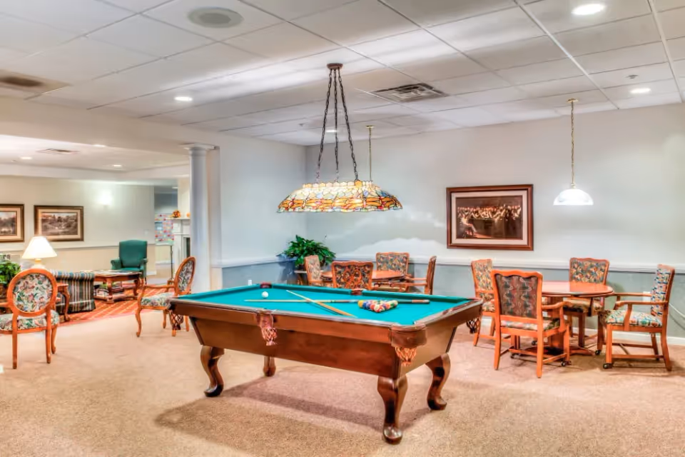 A well-lit common area in a senior living facility featuring a pool table with cues and balls arranged for a game. Surrounding the pool table are several wooden chairs with floral upholstery and small tables. The room has a beige carpet, white ceiling tiles with recessed lighting, and framed artwork on the walls. In the background, there is a seating area with armchairs and a lamp.