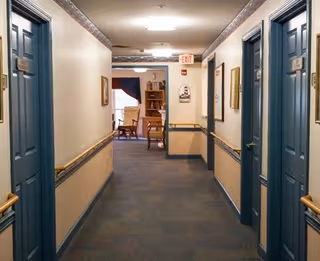 A hallway in a senior living facility with blue doors on both sides, beige walls with wooden handrails, and carpeted floor. At the end of the hallway, there is a room with chairs and a window with a curtain.