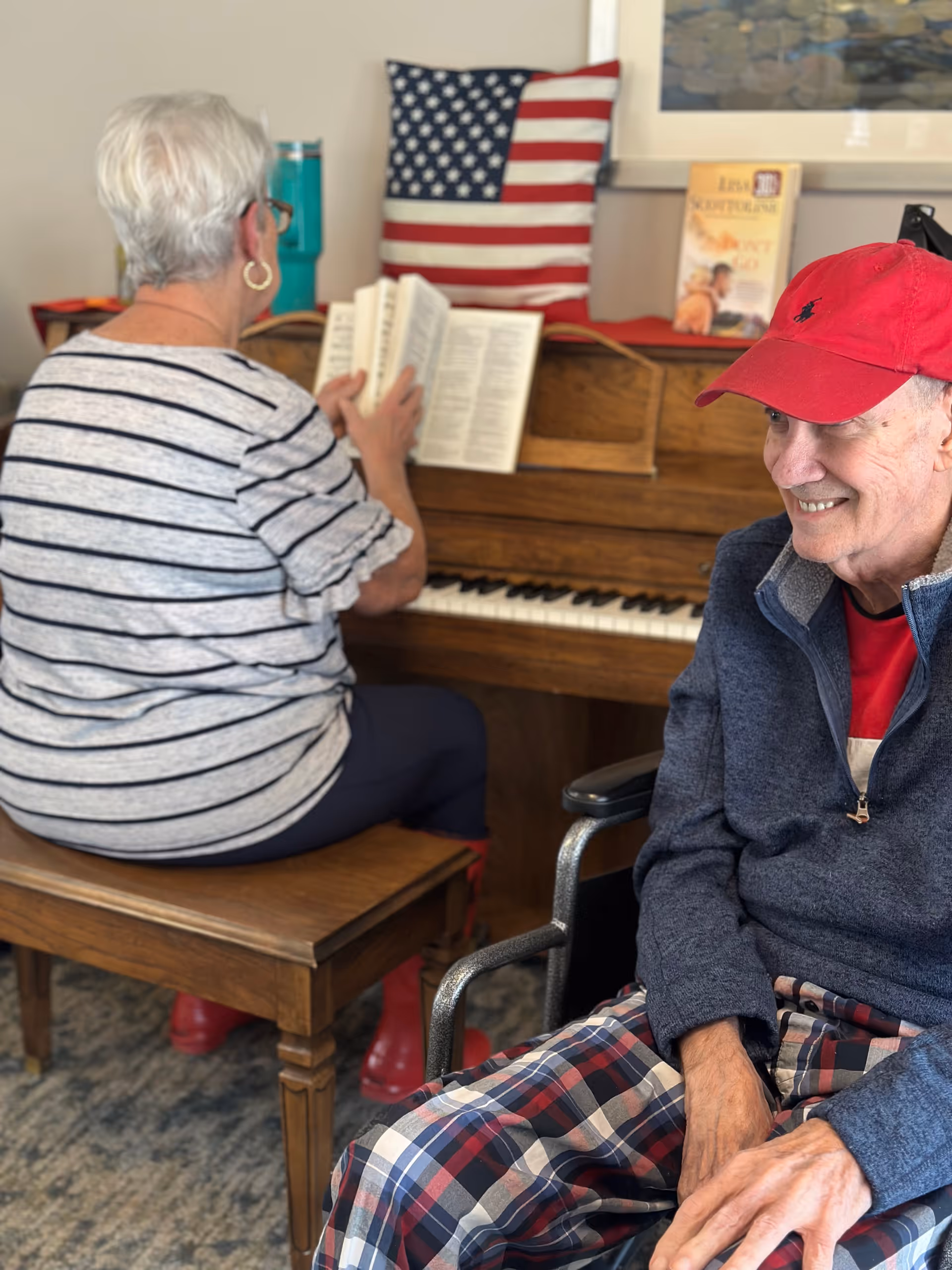 An elderly woman with short white hair and glasses is sitting on a wooden bench playing a piano, with an American flag pillow and a book on the piano. An elderly man wearing a red cap, blue jacket, and plaid pants is sitting in a wheelchair nearby, smiling.