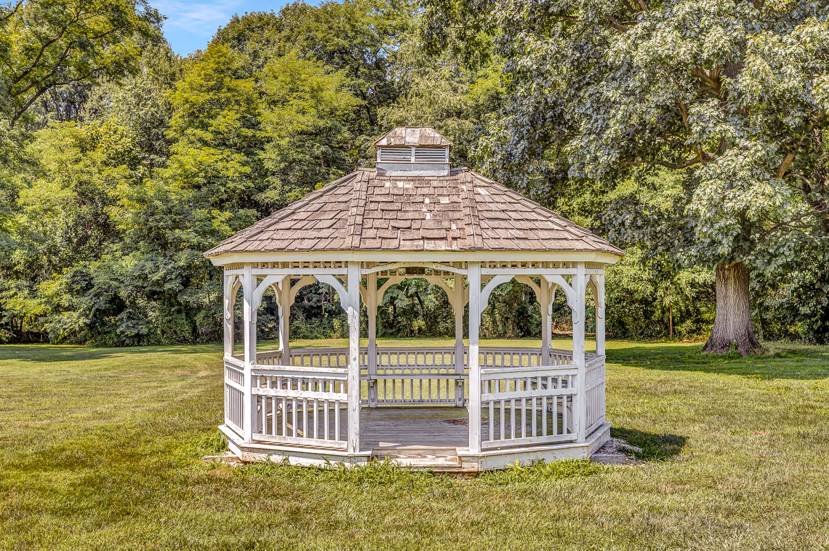 A white wooden gazebo with a shingled roof situated on a grassy lawn surrounded by trees and greenery under a clear blue sky.