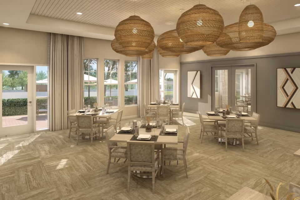 A bright dining room with multiple square tables set with plates, glasses, and napkins. The room features large windows with beige curtains allowing natural light to enter. There are several woven pendant light fixtures hanging from the ceiling. The walls are decorated with modern geometric artwork, and there is a door leading outside to a patio area with umbrellas and greenery.