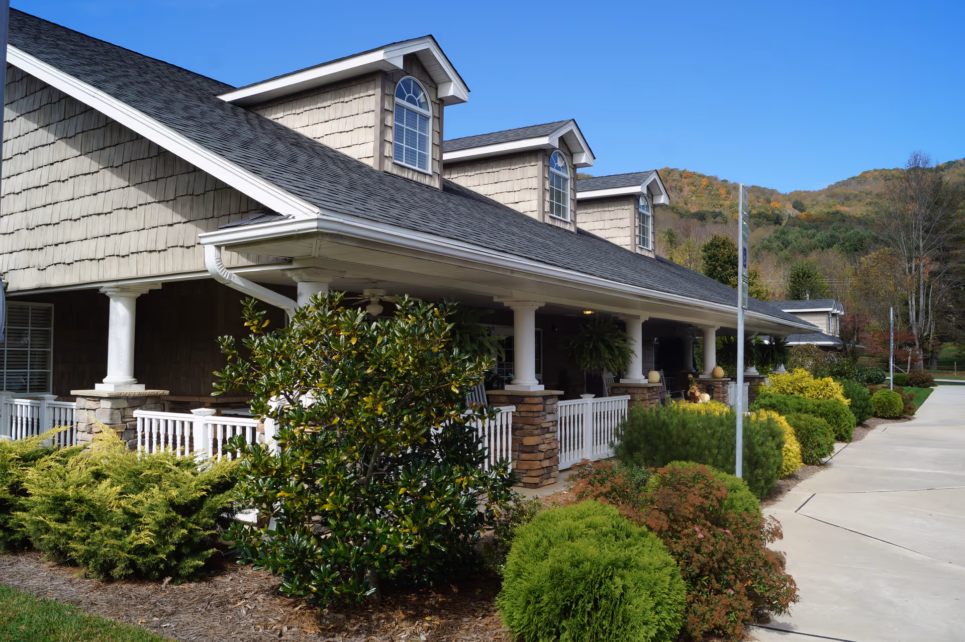 Exterior view of a senior living facility building with a covered porch supported by white columns and stone bases. The building has a shingled roof with dormer windows and is surrounded by well-maintained bushes and greenery. A paved walkway runs alongside the building, and there are hills with trees in the background under a clear blue sky.