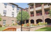 Courtyard with landscaping, a bench, and the brick façade of a multi-story assisted living building with balconies and arched walkways.