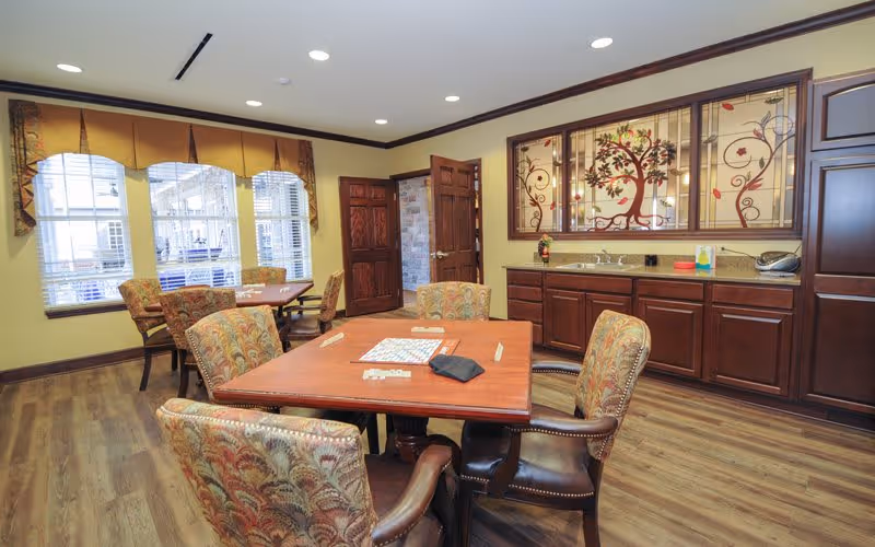 A cozy interior room with wooden flooring and yellow walls featuring a table with a board game and four upholstered chairs around it. In the background, there is another table with chairs near windows with blinds and valances. A wooden door is open, and a decorative stained glass panel with a tree design is mounted above a wooden cabinet with a countertop.