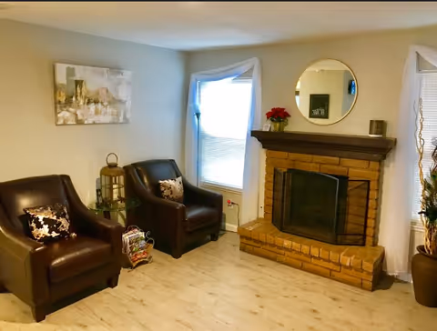 A cozy living room with two dark brown leather armchairs, each with a decorative pillow. Between the chairs is a small glass side table with a lantern and magazines underneath. The room features a brick fireplace with a wooden mantle, decorated with a round mirror, a small plant, and a candle. There are two windows with white curtains on either side of the fireplace, and light-colored wooden flooring.