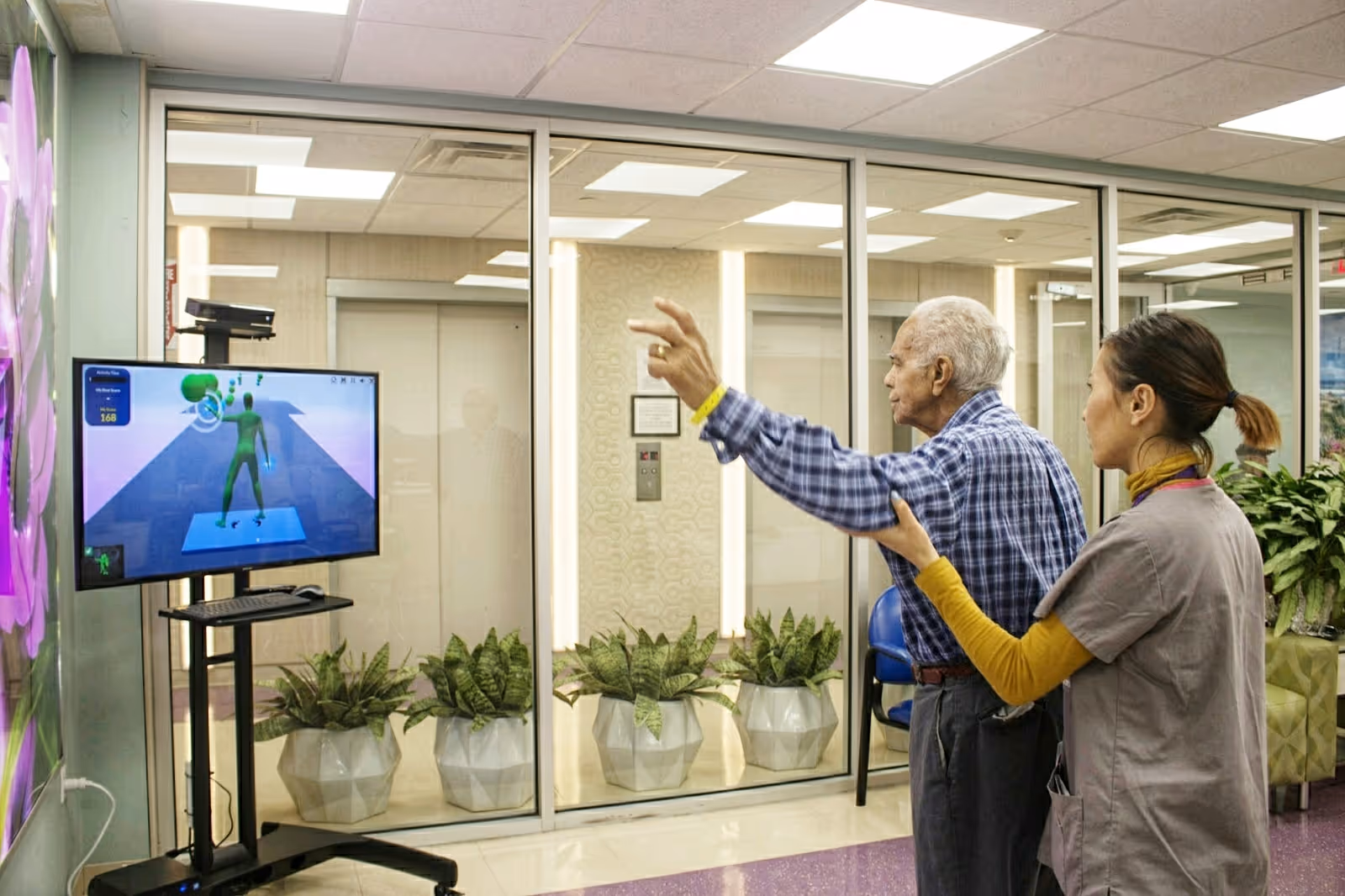 An elderly man wearing a blue checkered shirt is guided by a female caregiver in gray scrubs as he interacts with a motion-sensing game displayed on a TV screen mounted on a stand. The room has glass walls with potted plants lined up against them and bright overhead lighting.