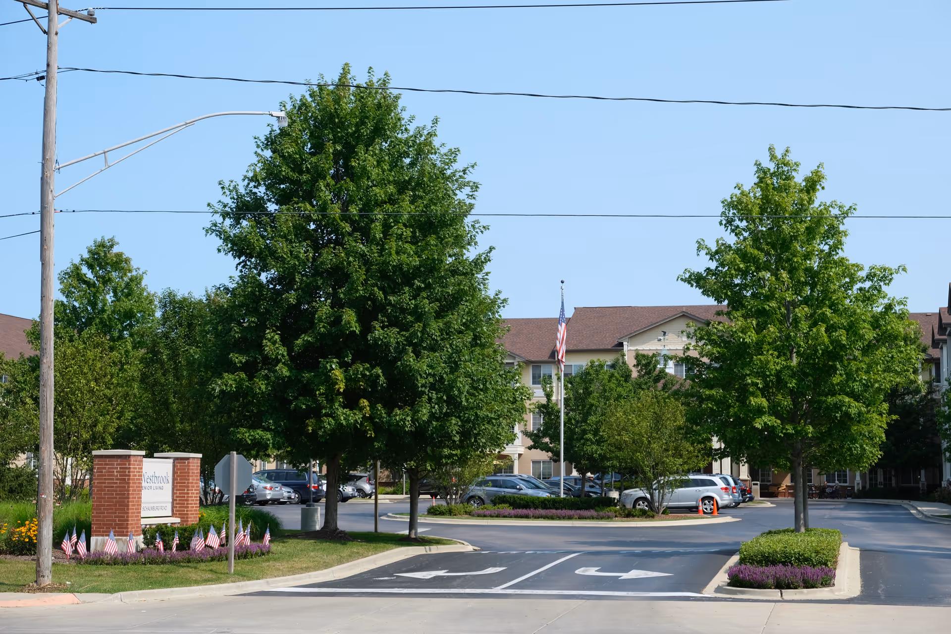 Exterior view of Westbrook Senior Living facility showing a driveway entrance with directional arrows, a brick sign with the facility name, several trees, parked cars, and an American flag on a flagpole under a clear blue sky.