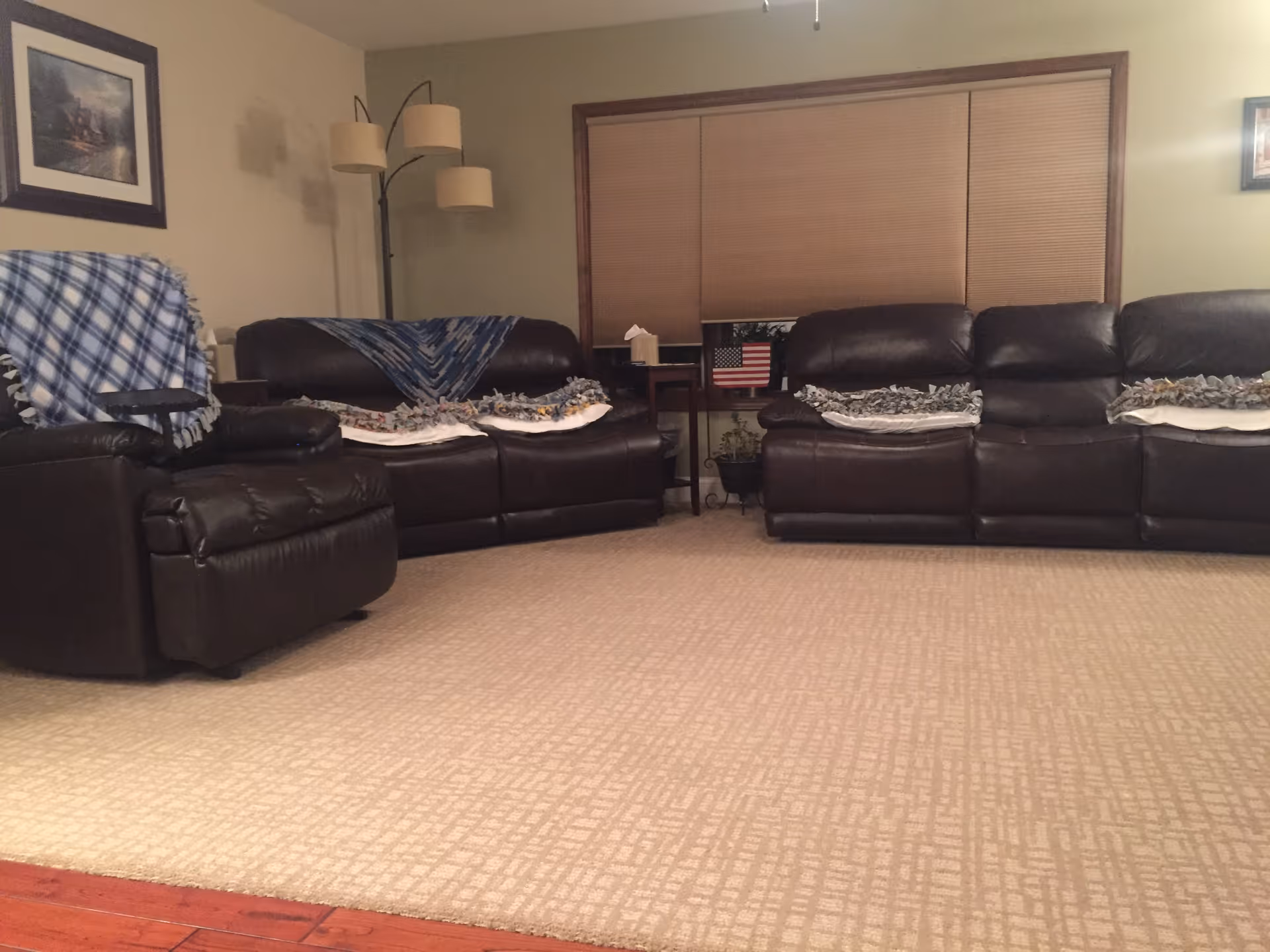 A cozy living room with two dark brown leather sofas and a matching recliner chair. Each sofa has several folded blankets draped over the backrests. There is a tall floor lamp with three lampshades in the corner, a small side table between the sofas with a tissue box and a small American flag decoration. The window behind the sofas has closed blinds, and the walls are painted beige with framed pictures hanging.