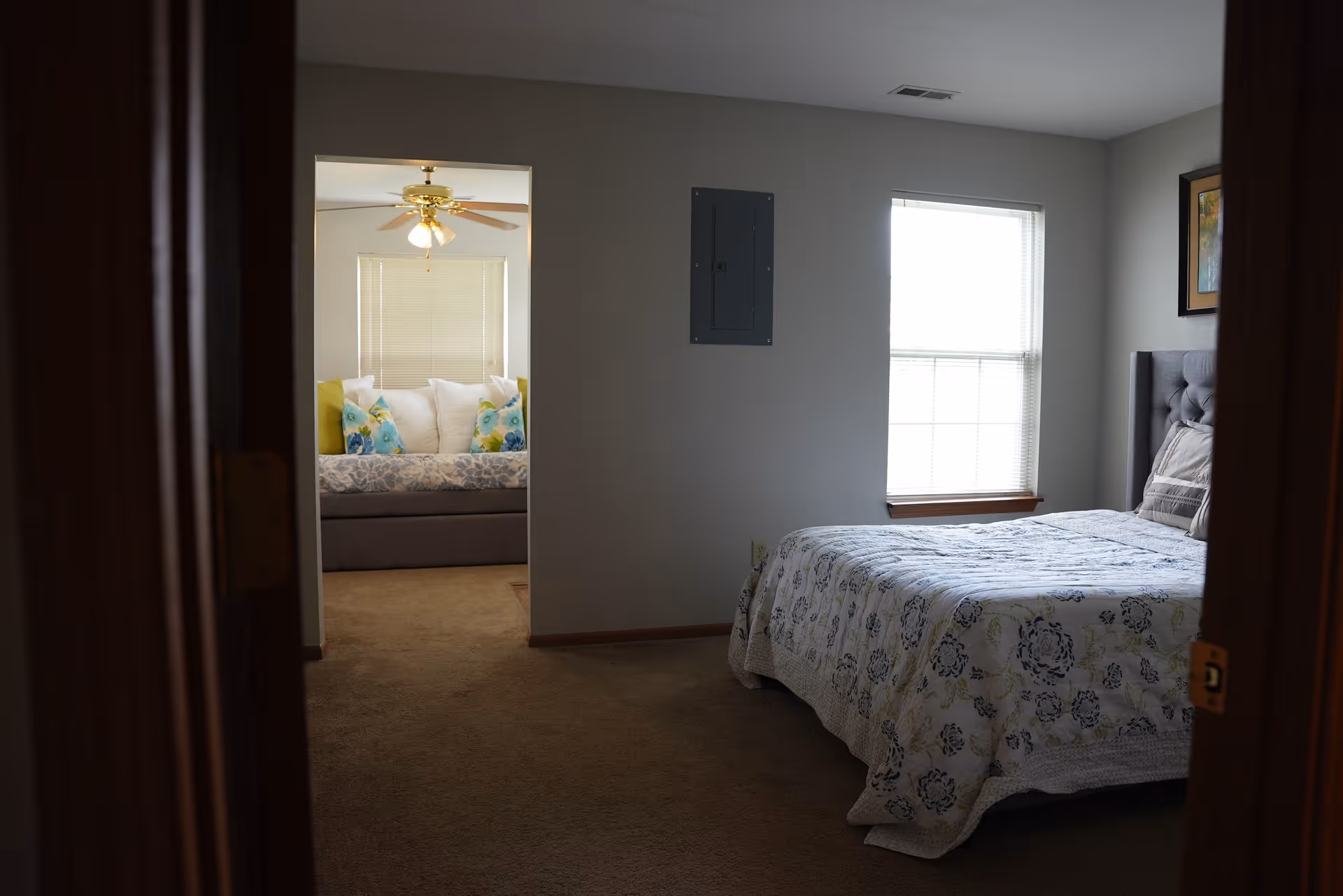 View into a softly lit bedroom with a floral quilted bed on the right and a cozy seating nook with colorful pillows visible through an open doorway.