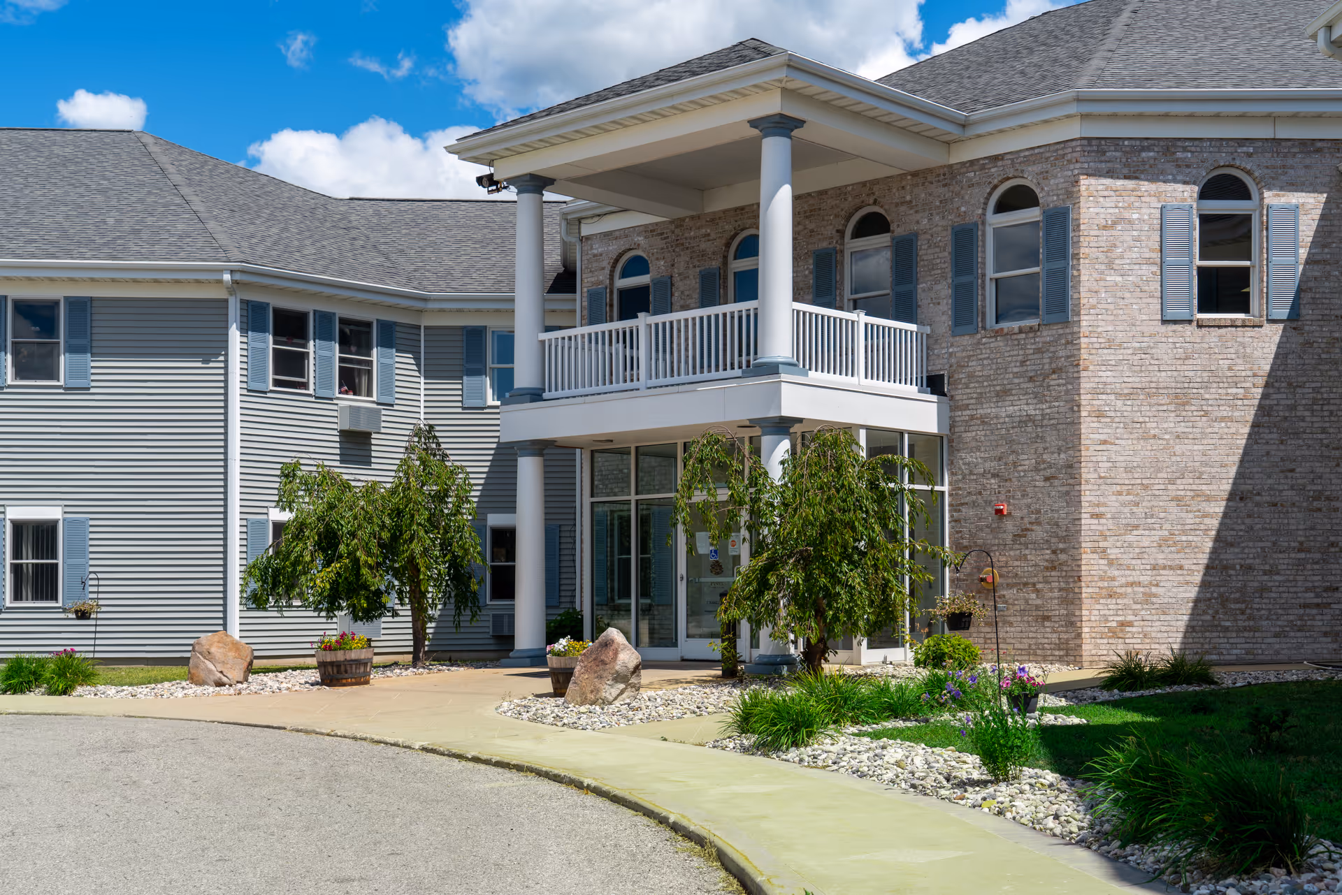 Exterior view of a senior living facility building with a covered entrance supported by columns, surrounded by small trees, plants, and rocks under a blue sky with some clouds.