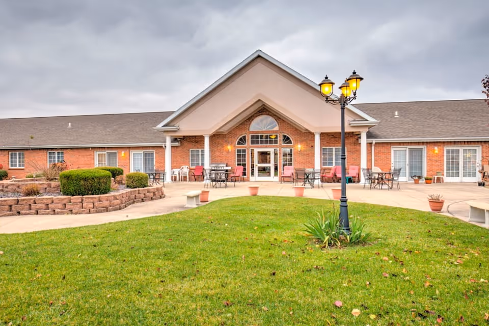 Exterior view of a single-story brick building with a peaked roof and large central entrance. The entrance area has glass doors and windows, outdoor seating with tables and chairs, and a lamppost with three lit lanterns on a grassy lawn in the foreground. The sky is overcast.