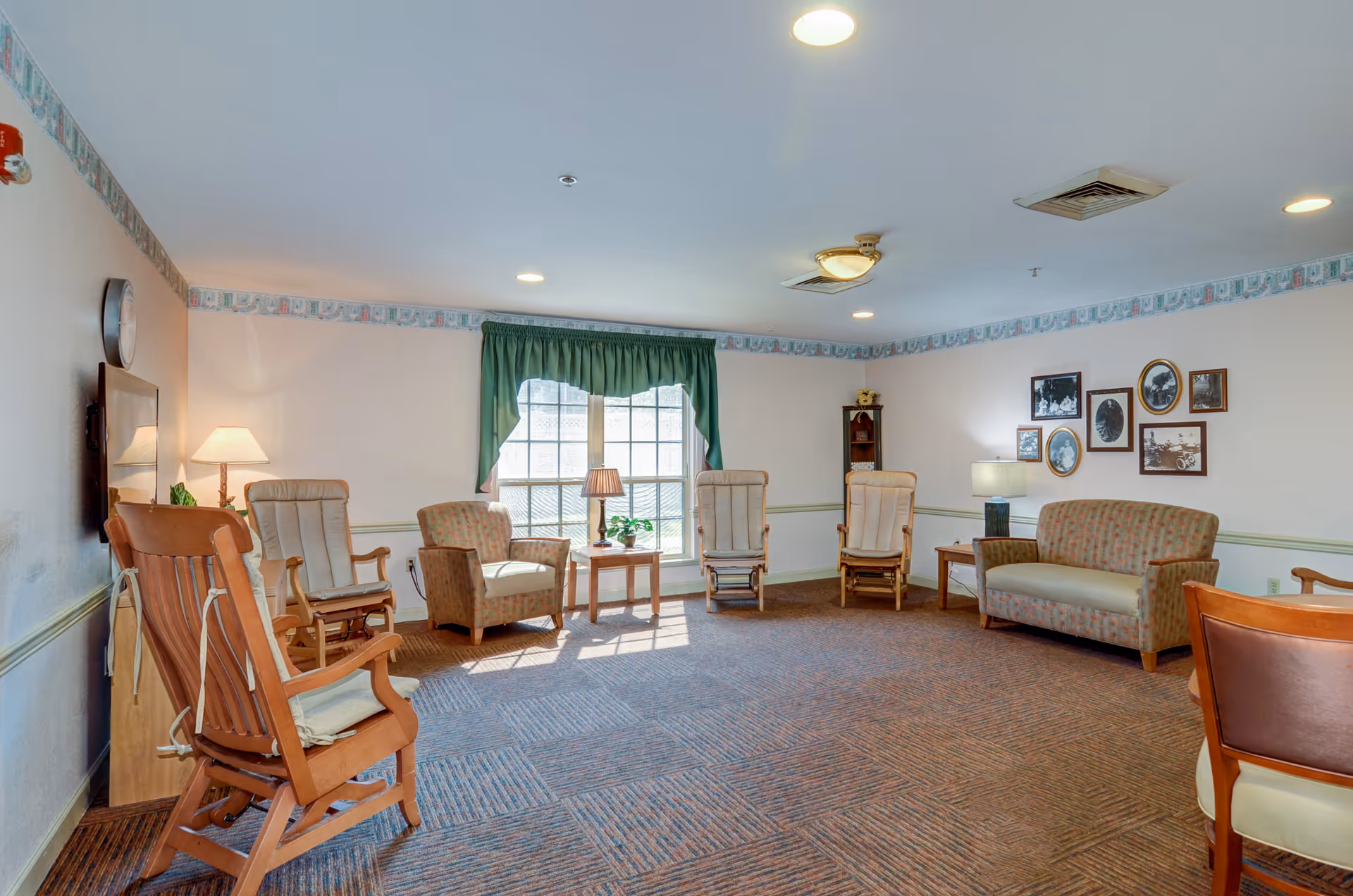 A senior living facility common room with several wooden rocking chairs and cushioned armchairs arranged around the perimeter. There is a window with green curtains letting in natural light, a few side tables with lamps and plants, and framed black-and-white photos on the wall. The room has carpeted flooring and a ceiling with recessed lighting.