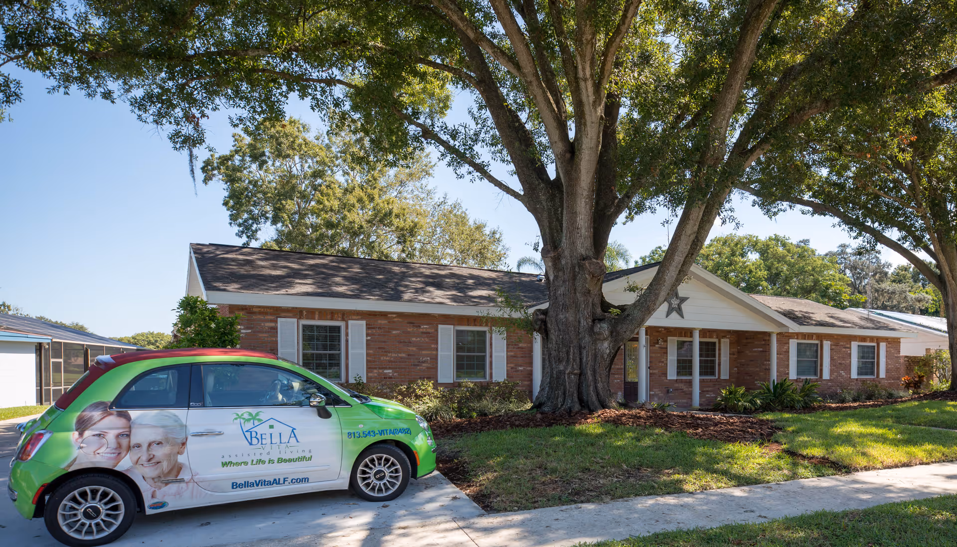 A single-story brick building with white shutters and a large tree in front, next to a small green car with Bella Vita ALF branding parked on the driveway.