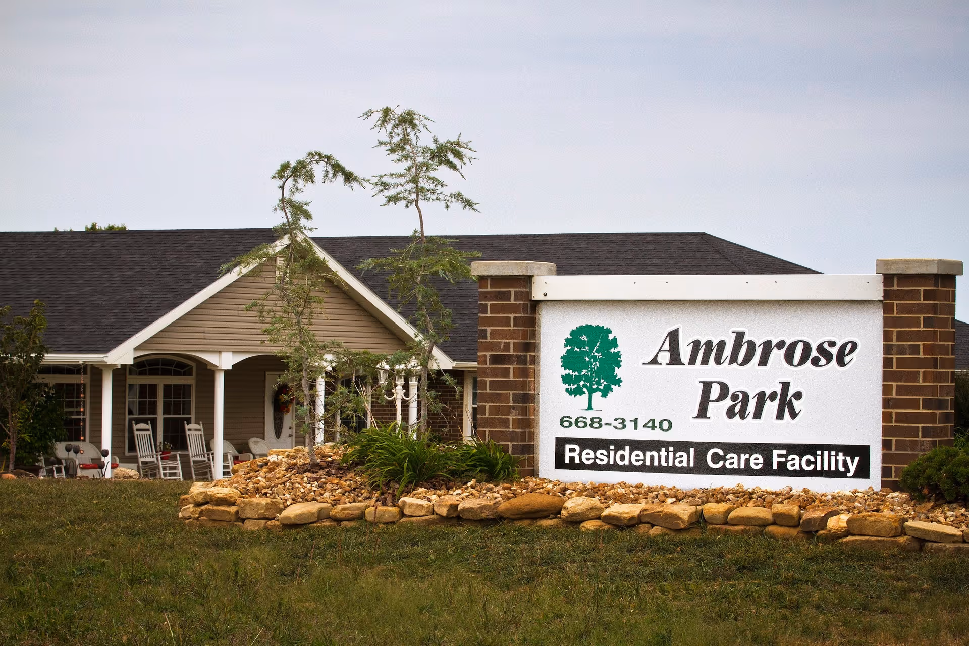 A large sign reading "Ambrose Park Residential Care Facility" in front of a single-story building with a porch and rocking chairs.