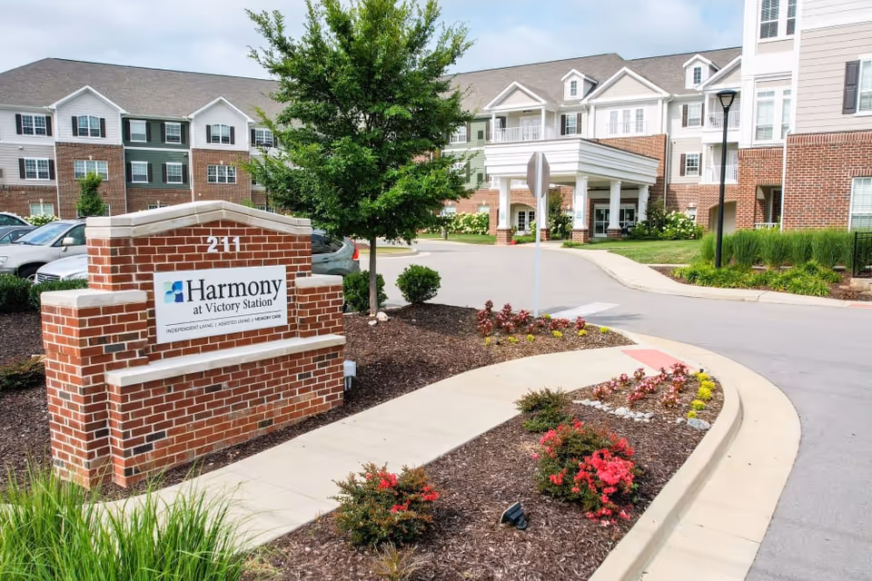 Exterior view of Harmony at Victory Station senior living facility showing a brick sign with the facility name, landscaped flower beds, a tree, and the building entrance with a covered drop-off area.