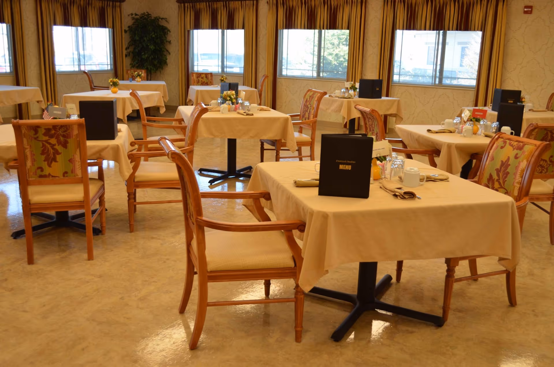 A dining room with multiple tables covered in beige tablecloths, each set with menus, cups, glasses, napkins, and small flower arrangements. The chairs have wooden frames with patterned upholstery. Large windows with yellow curtains allow natural light to fill the room.