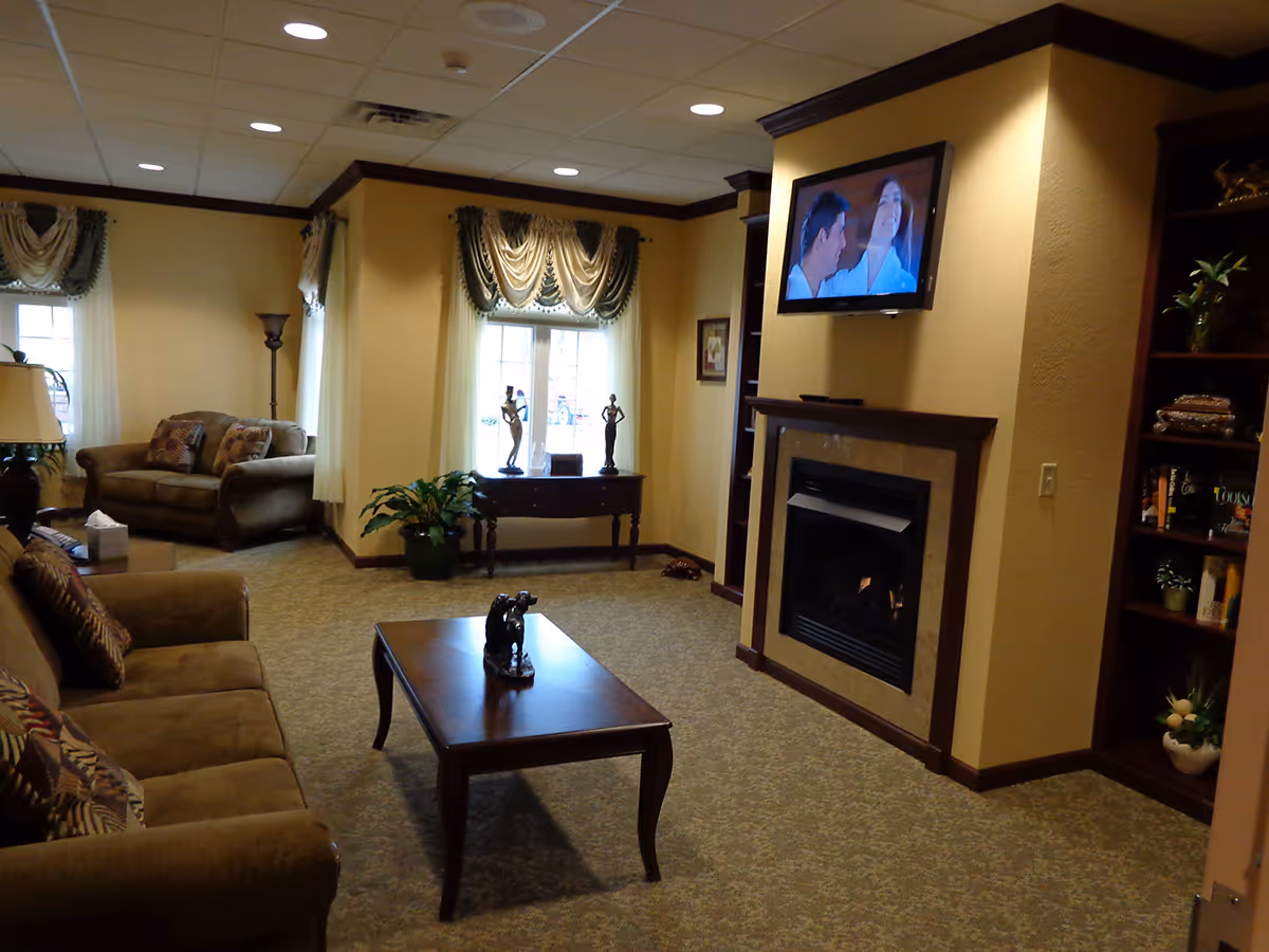A cozy living room area with beige walls and carpet, featuring two sofas with patterned cushions, a wooden coffee table with a small sculpture, a fireplace with a mounted flat-screen TV above it, a wooden bookshelf with books and plants, and a window with decorative curtains letting in natural light.