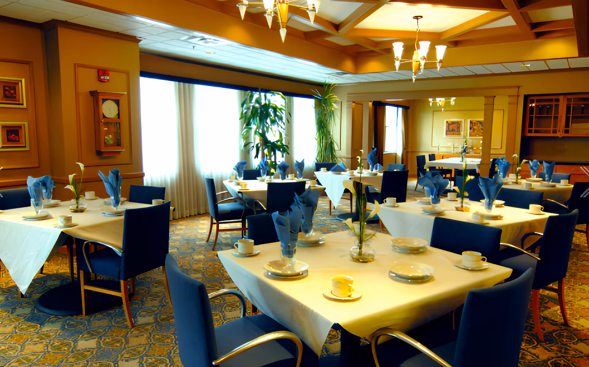 A well-lit dining room with multiple tables covered in white tablecloths, each set with blue napkins, white plates, cups, and small vases with flowers. The room has blue upholstered chairs, large windows with sheer curtains, decorative ceiling lights, and framed artwork on the walls.