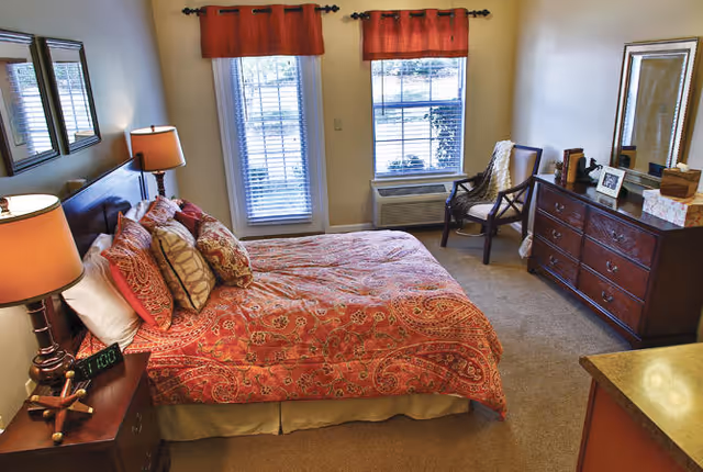 Bright bedroom with a red-patterned bed, bedside tables and lamps, a dresser with mirror, a chair, and two windows with red valances.