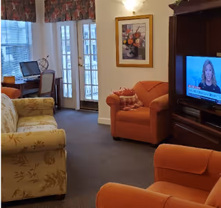 A cozy living room with floral and orange upholstered armchairs and a sofa. A wooden entertainment center holds a television showing a news program. There is a framed floral painting on the wall and a window with floral valances letting in natural light. A desk with a chair and a computer is visible near a set of glass-paneled double doors.