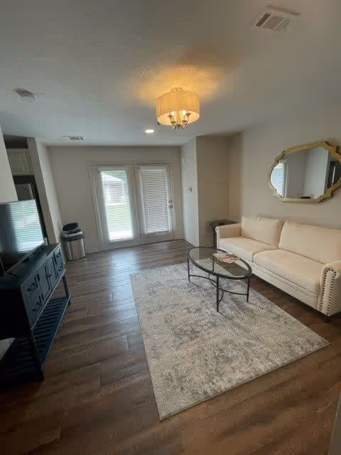 A bright living room with wooden flooring, a beige sofa, a glass coffee table on a patterned rug, a wall-mounted decorative mirror, a TV on a black stand, and double glass doors leading outside.