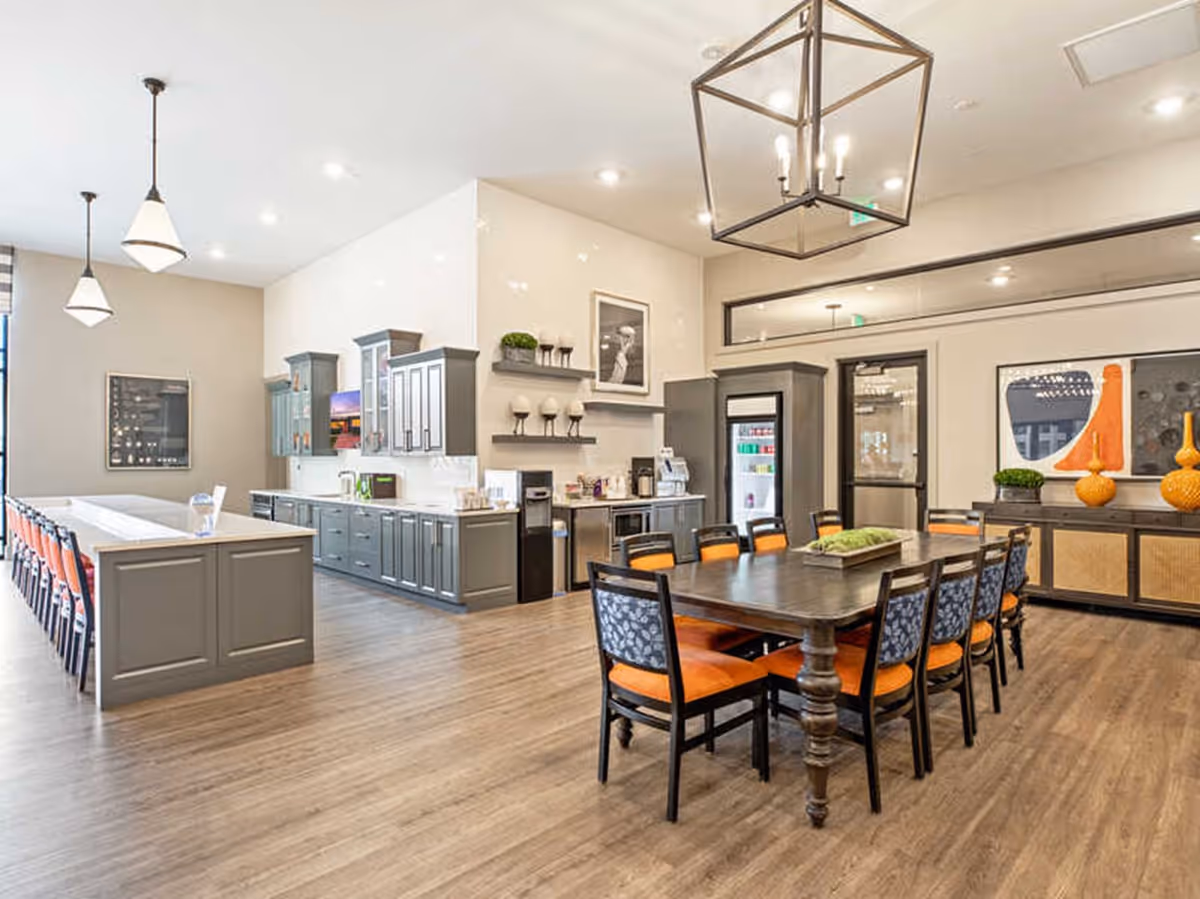 Open communal dining and kitchen area with a long island and a large dining table surrounded by orange-cushioned chairs.