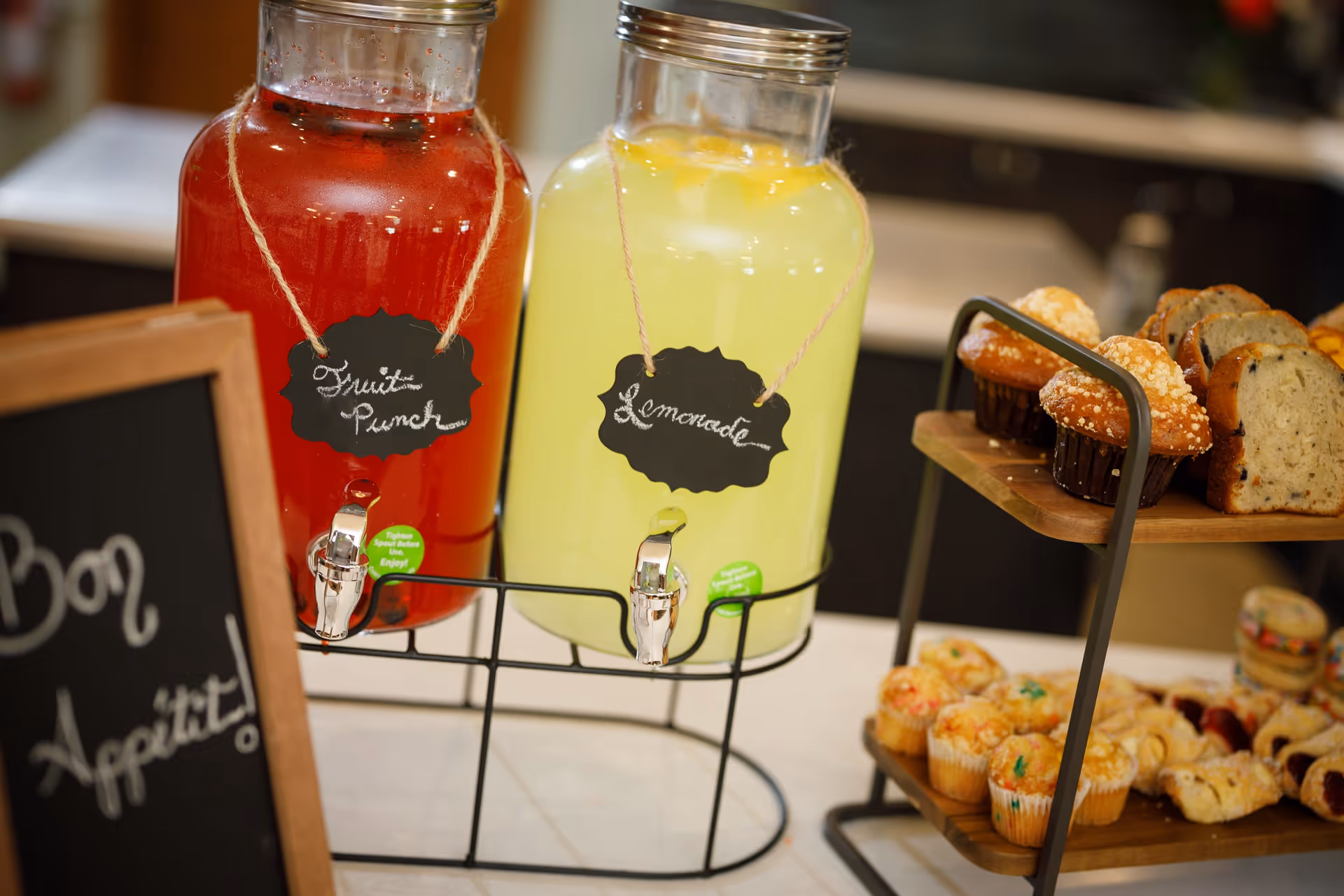 Two large glass beverage dispensers on a metal stand, one filled with red fruit punch and the other with yellow lemonade, each labeled with a small chalkboard sign. Next to the dispensers is a two-tiered wooden tray holding muffins, bread slices, and assorted pastries. A chalkboard sign with the words 'Bon Appetit!' is partially visible on the left side.