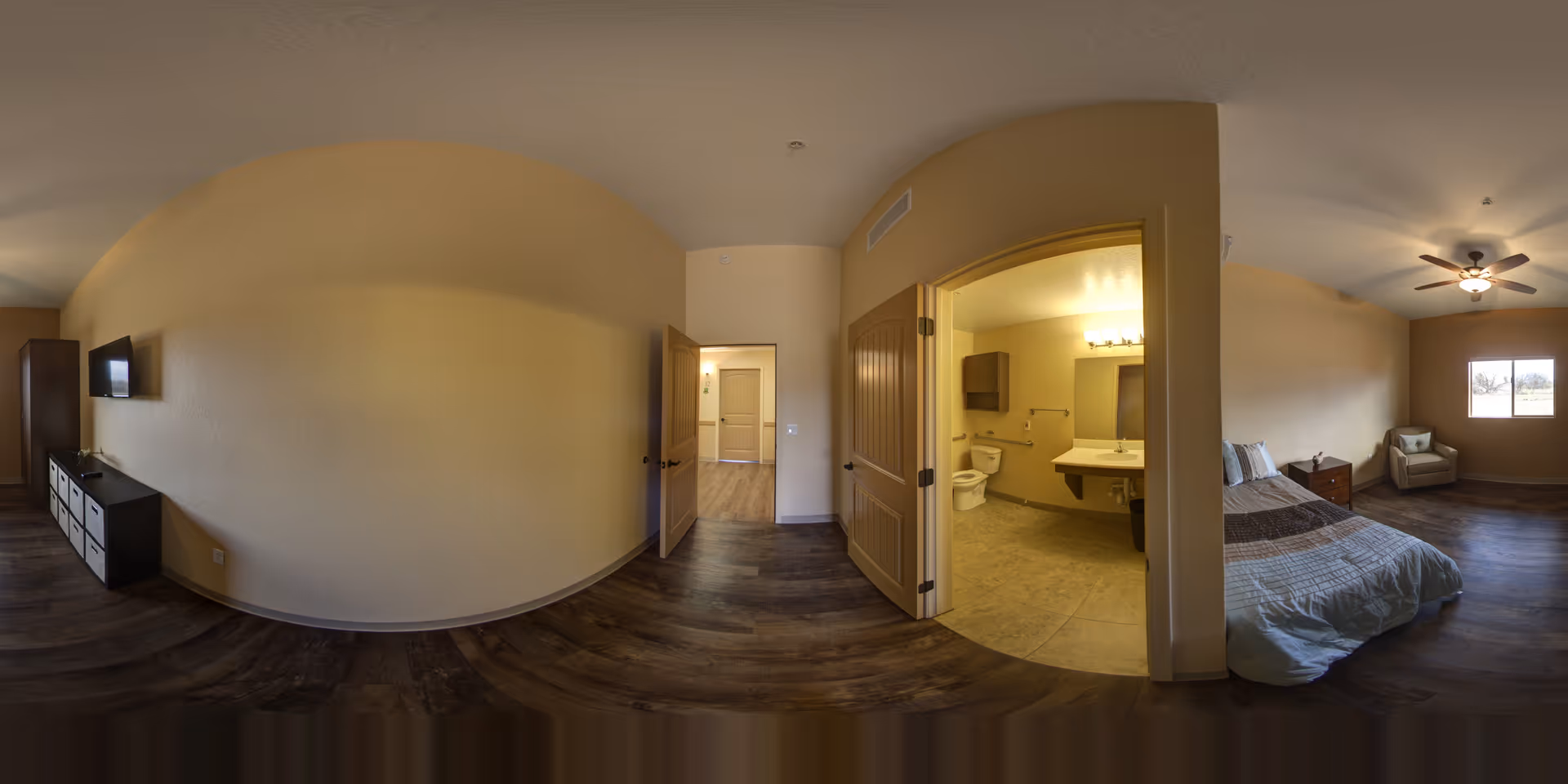 A spacious assisted living apartment interior with a bedroom and an attached bathroom. The bedroom features a bed with pillows and a blanket, a nightstand, an armchair near a window, and a ceiling fan with a light. The bathroom has a toilet, a sink with a large mirror, and a cabinet. The flooring is dark wood laminate, and the walls are painted beige. There is a TV mounted on the wall opposite the bed and a dresser below it.