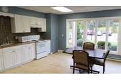 A bright kitchen area with white cabinets, a white stove, and a countertop. There is a dining table with four chairs near large windows that look out onto a green outdoor space with bushes and parked cars.