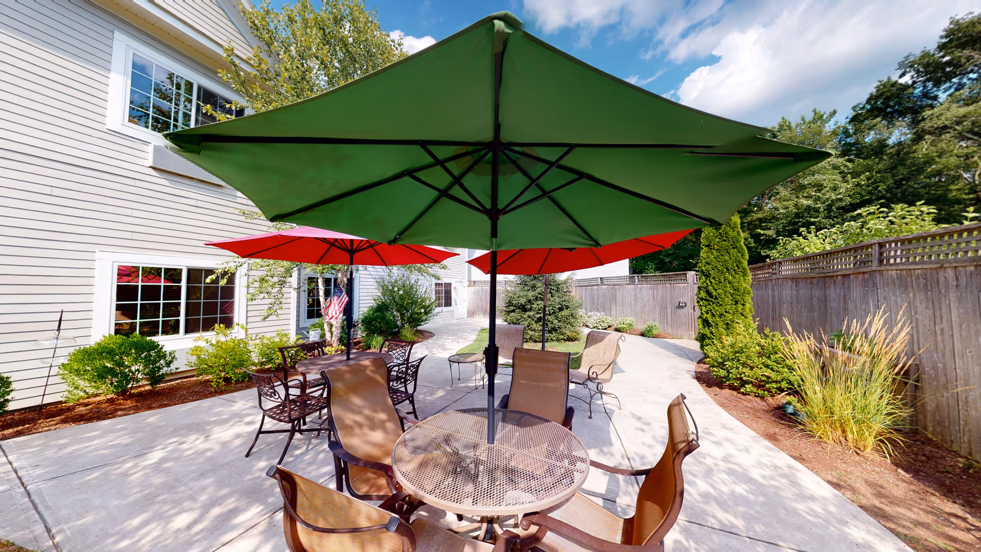 Outdoor patio area at The Enclave of Franklin with round metal tables and chairs under large green and red umbrellas, surrounded by greenery and a wooden fence.
