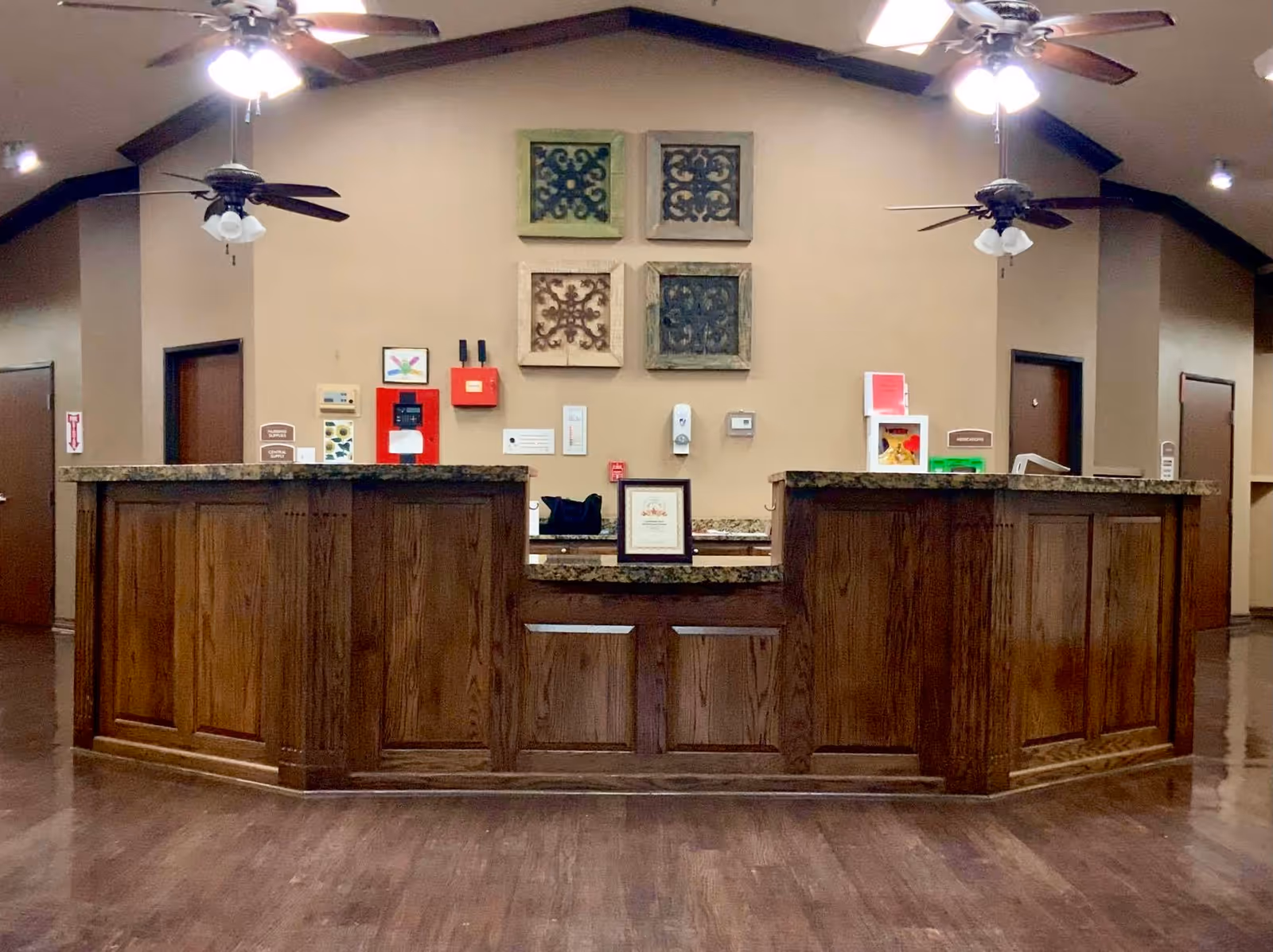 Reception desk area in a senior living facility with a wooden counter, decorative wall art above, two ceiling fans with lights, and doors on either side of the desk.