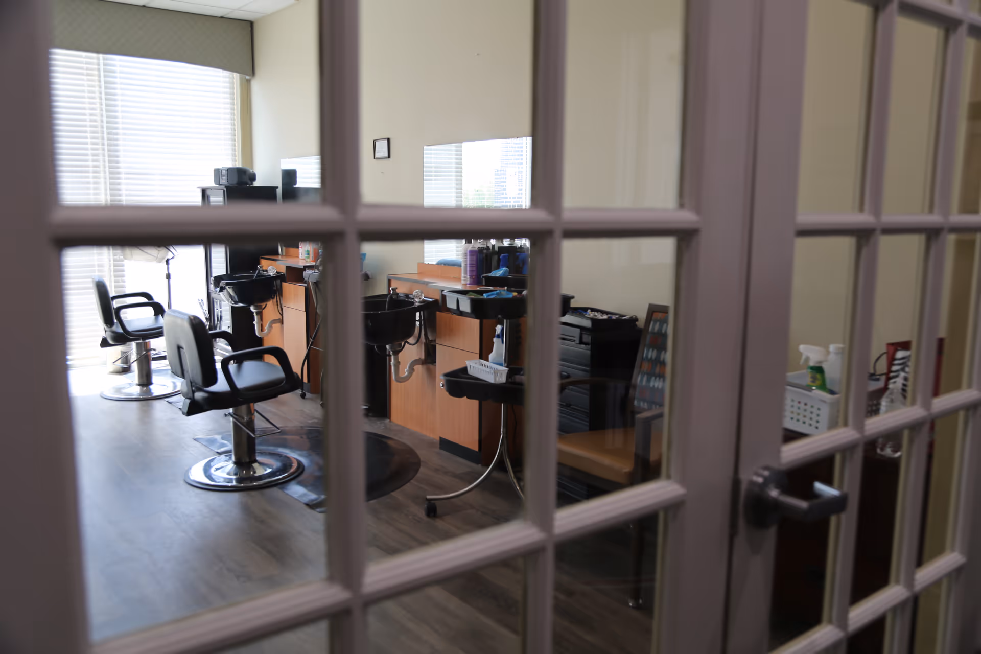 View through glass-paneled doors into a hair salon area with two black salon chairs, two black wash basins, wooden cabinetry, and various hair care products on the counters. The room has large windows with blinds allowing natural light to enter.