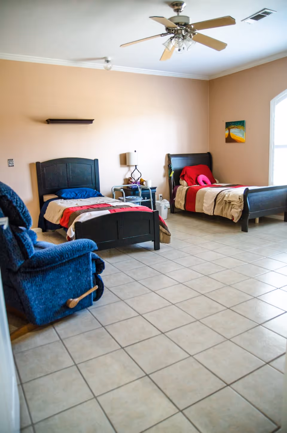 A senior living bedroom with two single beds, each with dark wooden frames and colorful bedding. There is a blue recliner chair in the foreground, a small nightstand with a lamp between the beds, a ceiling fan above, and a window letting in natural light. The floor is tiled and the walls are painted beige with a small painting on one wall.