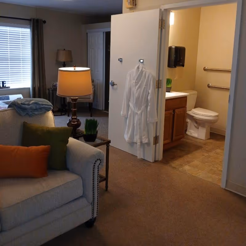 Interior view of a senior living facility room showing a beige couch with green and orange pillows, a side table with a lamp, and an open door leading to a bathroom with a white robe hanging on the door. The bathroom has a toilet, a sink with a wooden cabinet, and grab bars on the wall.