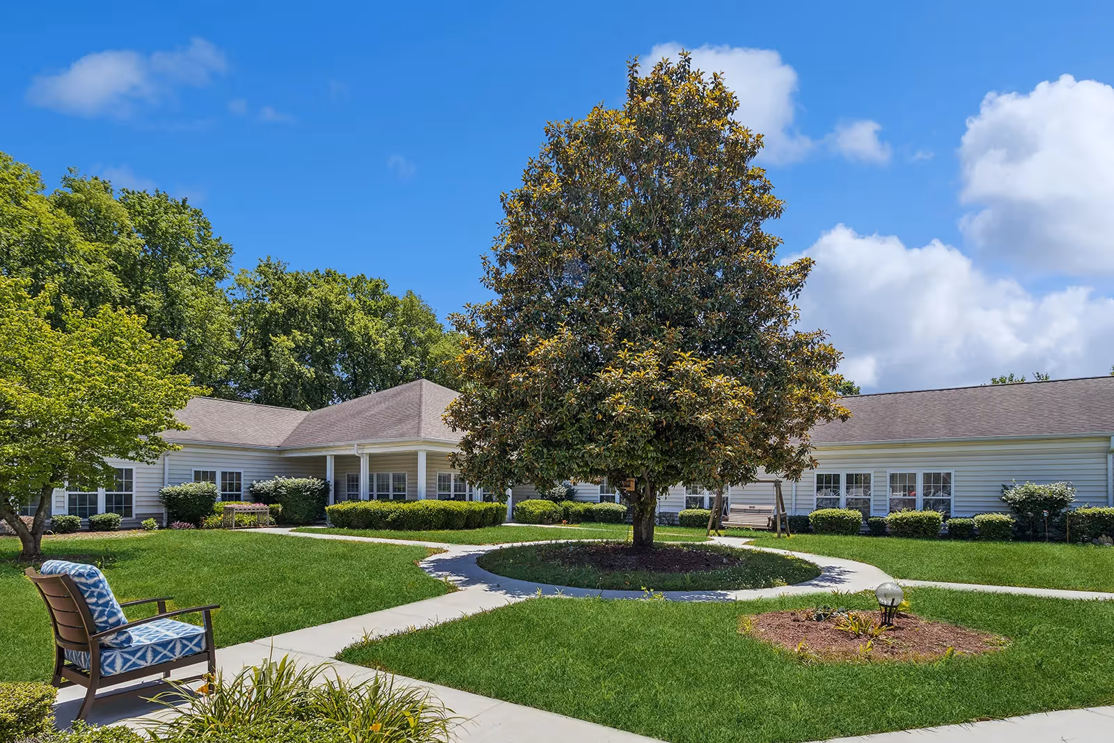 Courtyard with a large central tree, circular walkway, outdoor seating, and a single-story building under a blue sky.