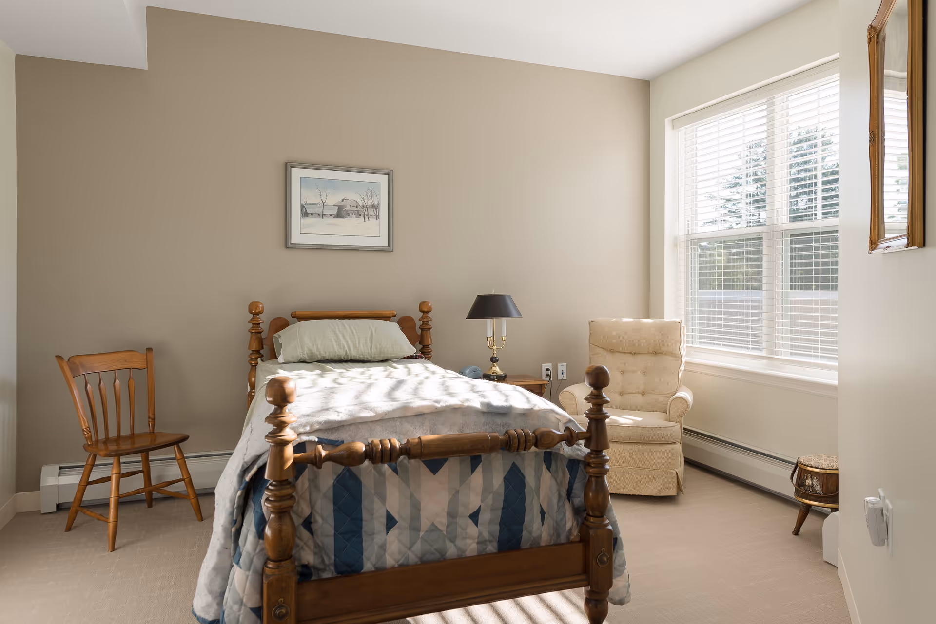 A bright bedroom with a wooden single bed covered with a patterned quilt and a pillow. There is a wooden chair to the left of the bed and a beige upholstered armchair near a large window with white blinds on the right. A small wooden nightstand with a black lamp is next to the bed. The walls are painted beige, and a framed picture hangs above the bed.