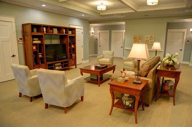A cozy senior living common area with beige carpet, light green walls, and white trim. The room features a wooden entertainment center with a TV, two patterned armchairs, a sofa, a wooden coffee table, and side tables with lamps and decorative items. There are multiple doors and soft ceiling lights providing warm illumination.