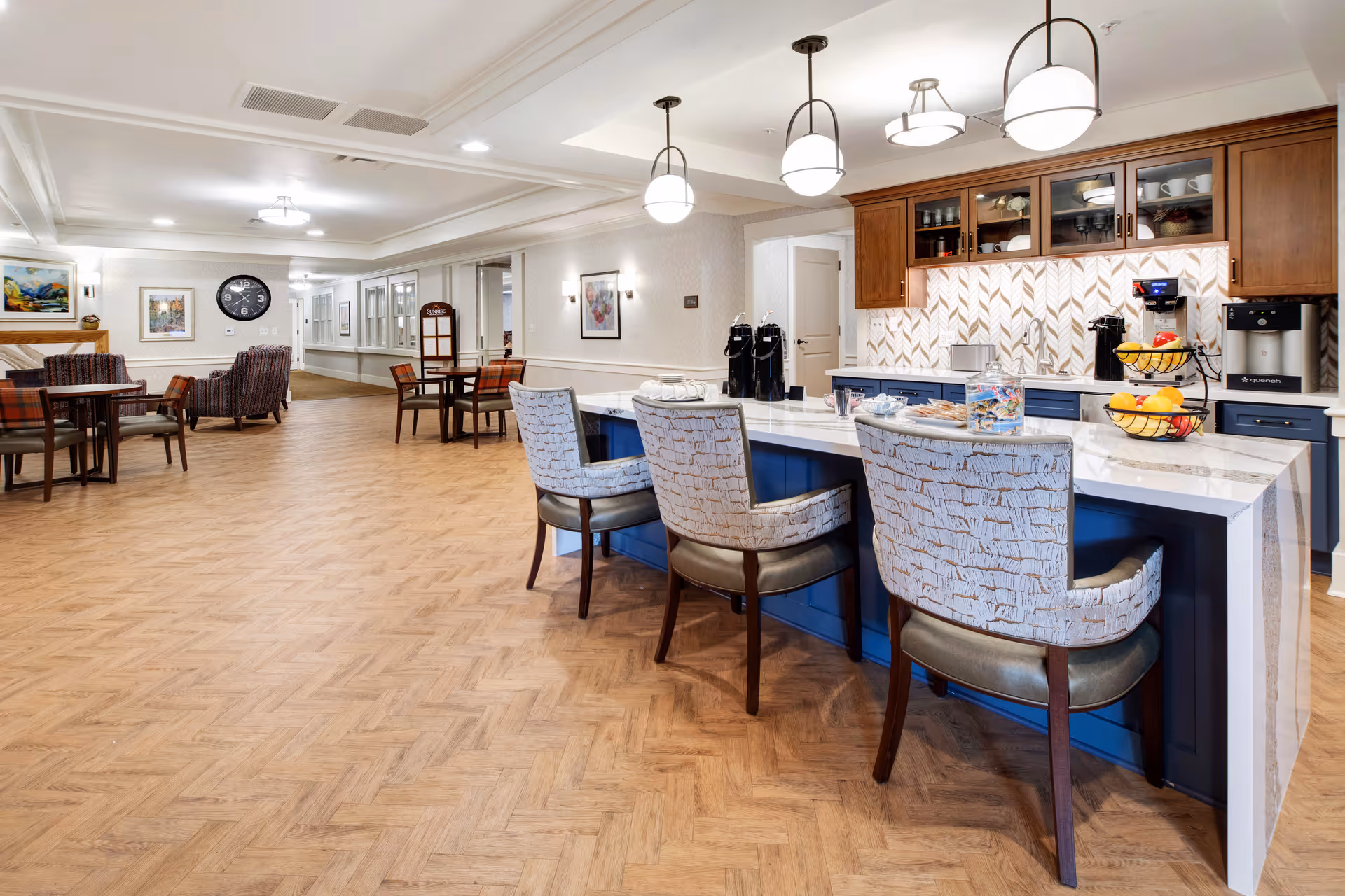A spacious and well-lit common area in a senior living facility featuring a kitchen island with three upholstered chairs, a coffee station with multiple coffee machines, and a fruit basket. The room has wooden flooring, pendant lights, and several tables and chairs arranged for dining or socializing. Artwork and a large clock decorate the walls.