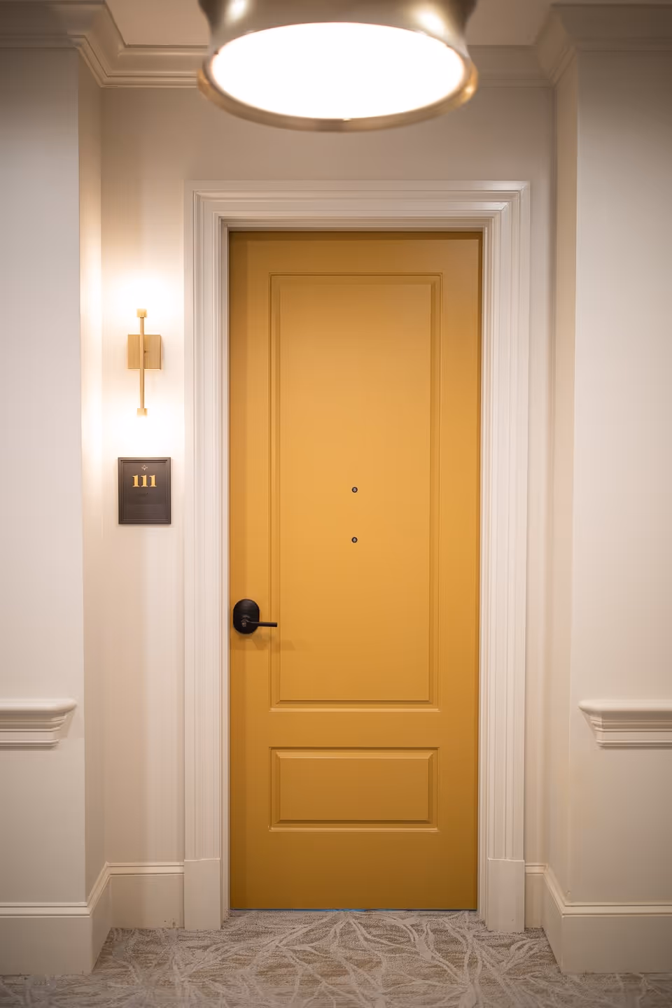 A mustard-yellow apartment door labeled 111 in a well-lit interior hallway with a wall sconce and overhead light.