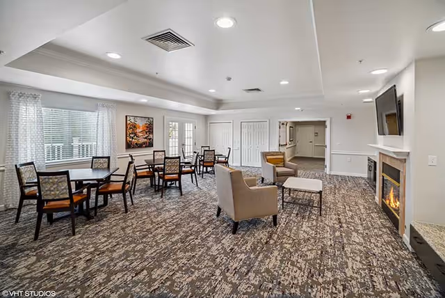 Spacious senior living common room with tables and chairs, lounge seating, a fireplace and wall-mounted TV.