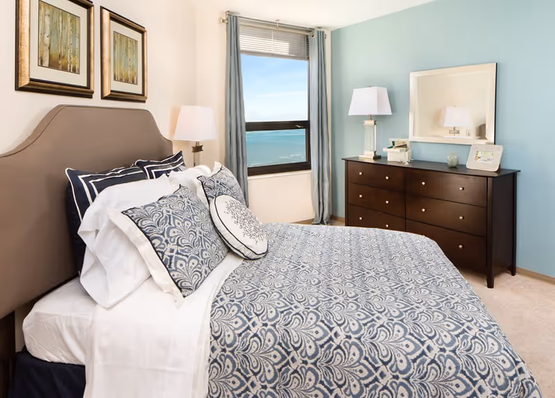 A neatly made bed with patterned blue and white bedding and multiple pillows in a bedroom. The room features a large window with blue curtains, a wooden dresser with a mirror, two table lamps, and two framed pictures on the wall above the bed.