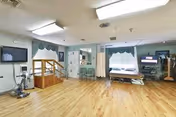 A spacious rehabilitation room with wooden flooring, exercise equipment including a stationary bike and parallel bars for walking practice, a hospital bed near the window with curtains, chairs, and a television mounted on the wall.