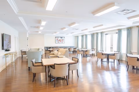 A bright and spacious dining room with multiple tables and chairs arranged neatly. Large windows with light curtains allow natural light to fill the room. The floor is wooden, and there is a wall-mounted TV on the left side along with a colorful painting on the far wall.