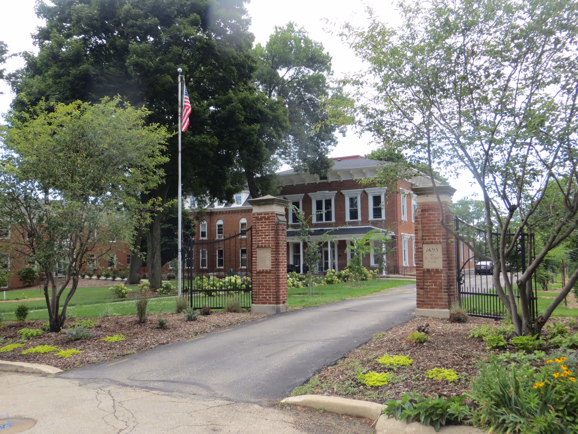 Front entrance of Mt. Pleasant Home showing a brick gate and driveway leading to a two-story brick building with a flagpole, trees, and landscaping.