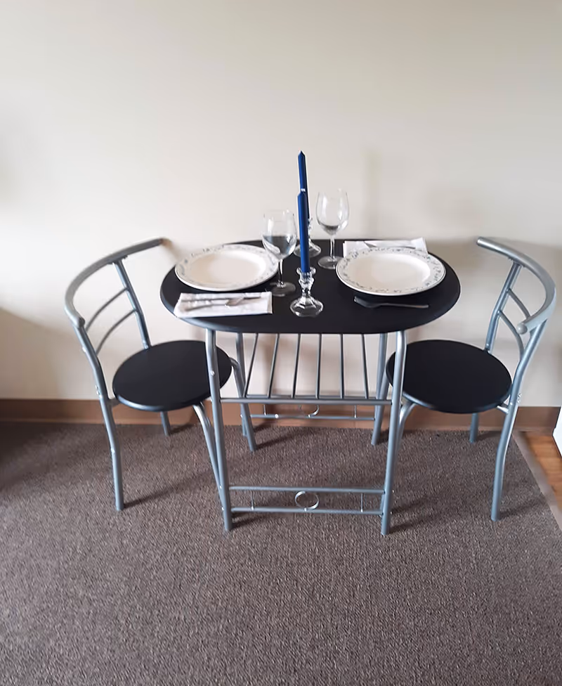 Small two-person dining table with two chairs set with plates, wine glasses, and a tall blue candle against a plain wall.