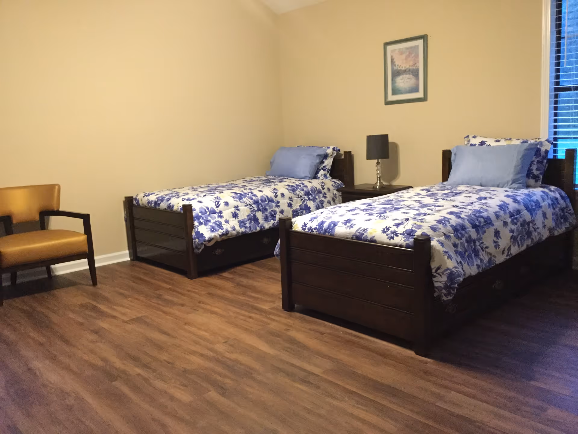 A bedroom with two single wooden beds, each made with white and blue floral bedding and blue pillows. Between the beds is a small nightstand with a lamp. There is a framed picture on the beige wall above the nightstand and a window with blinds on the right side. A brown chair is positioned on the left side of the room, which has wooden flooring.