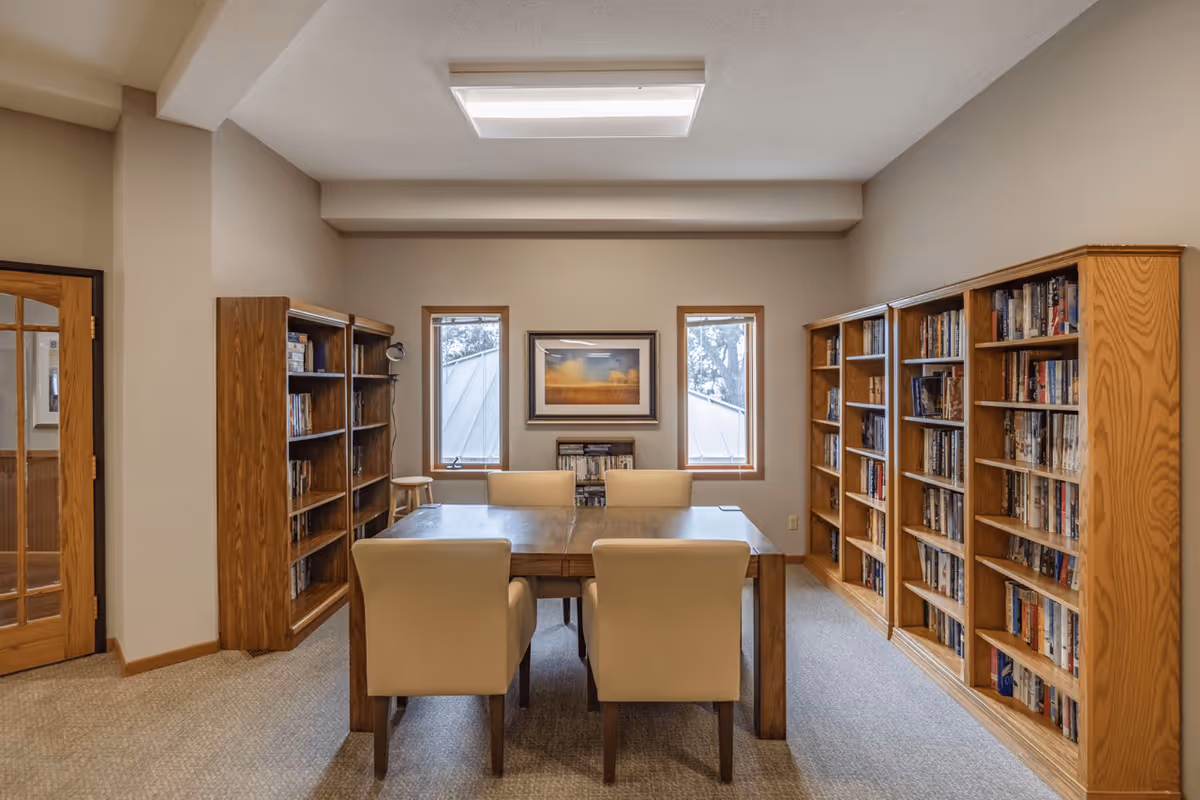 A cozy reading or meeting room with a wooden table surrounded by four beige upholstered chairs. The room has bookshelves filled with books on both sides, two narrow windows with wooden frames, and a framed picture hanging on the wall between the windows. The floor is carpeted, and there is a wooden door with glass panels on the left side.