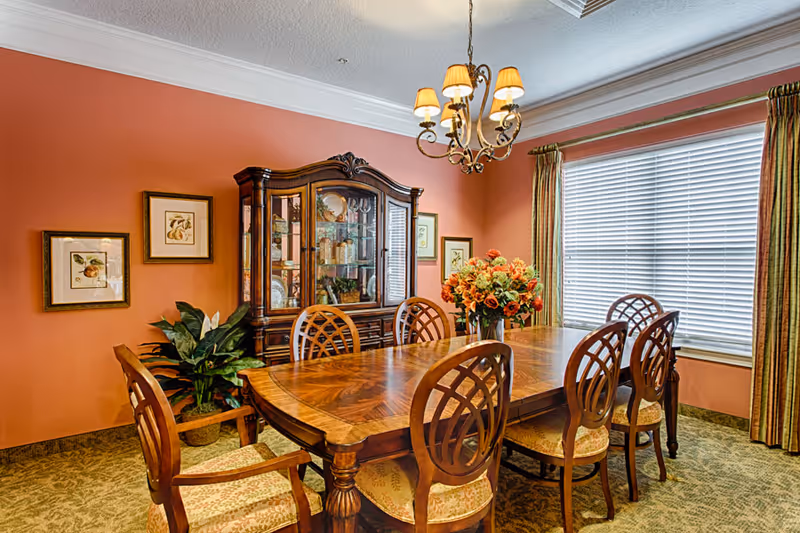 A dining room with a wooden dining table surrounded by six matching chairs with cushioned seats. A floral centerpiece is placed on the table. The walls are painted a warm orange color and decorated with framed artwork. A wooden china cabinet with glass doors displays dishes and glassware. A chandelier with five lampshades hangs above the table. A large window with closed blinds and curtains is on the right side of the room. There is a green potted plant in the corner.