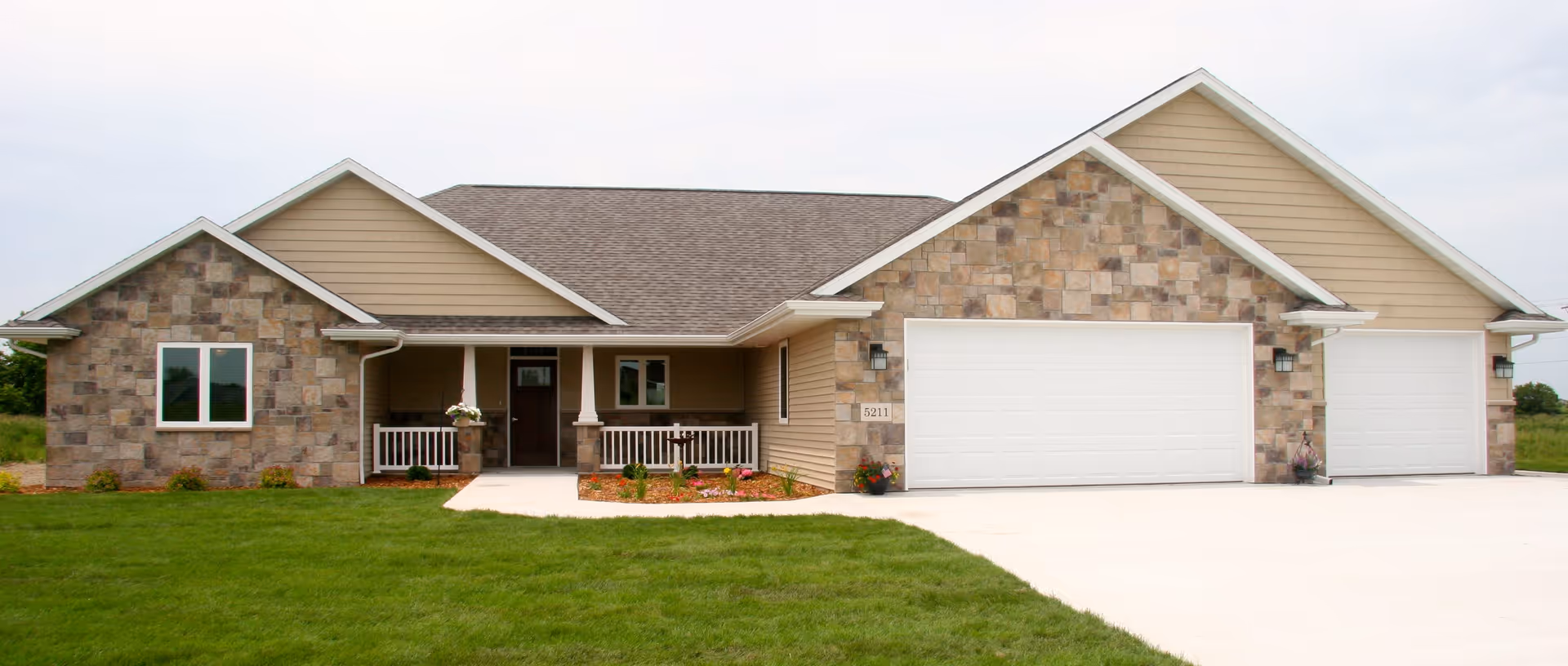 Front exterior view of a single-story house with a stone and beige siding facade, a covered porch with white railings, a dark brown front door, and a three-car garage with white doors. The house is surrounded by a green lawn and a concrete driveway.