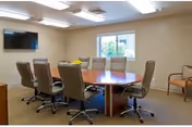 A conference room with a large oval wooden table surrounded by eight office chairs on wheels. There is a wall-mounted flat screen TV on the left wall and a window with a view of greenery on the far wall. The room has beige walls and carpeted flooring with ceiling lights.